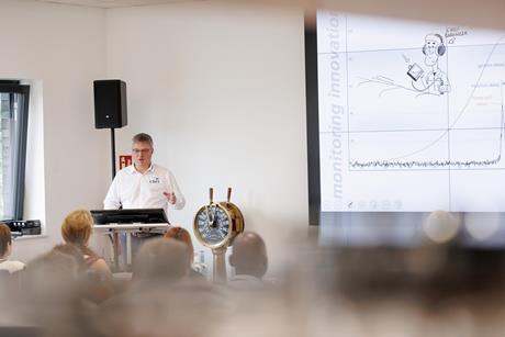A trainer stands by a white board and a ship's wheel training people